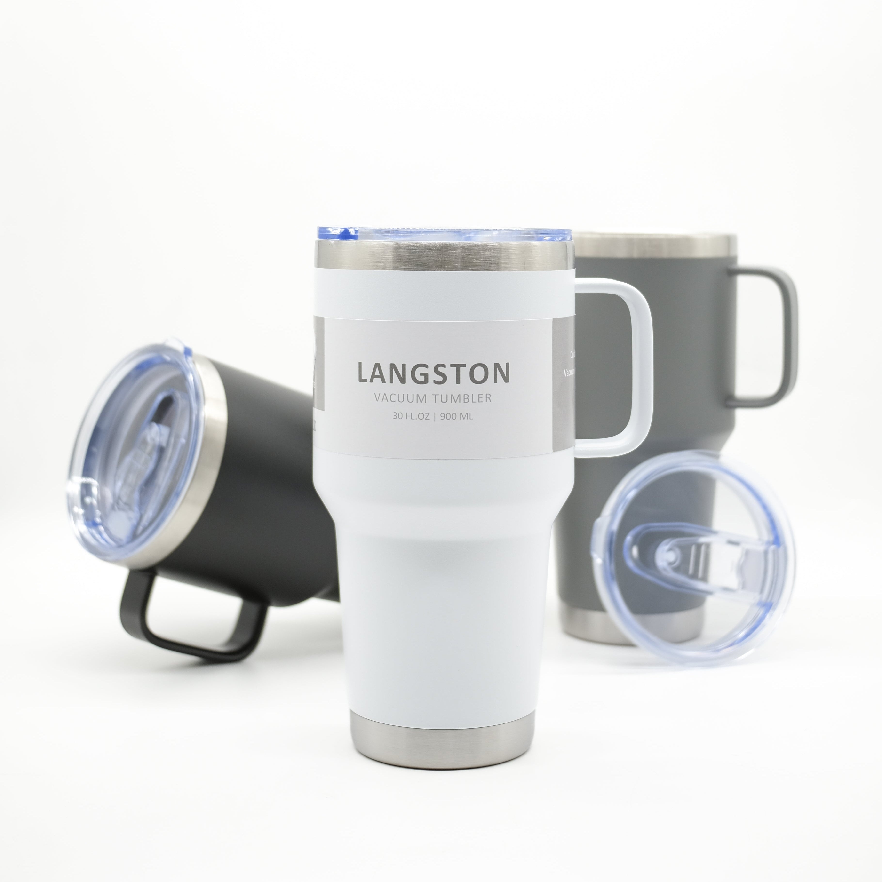 A white Langston vacuum insulated travel tumbler with a handle and clear lid, shown in front of a dark gray tumbler and a black tumbler with lid off on a white background.
