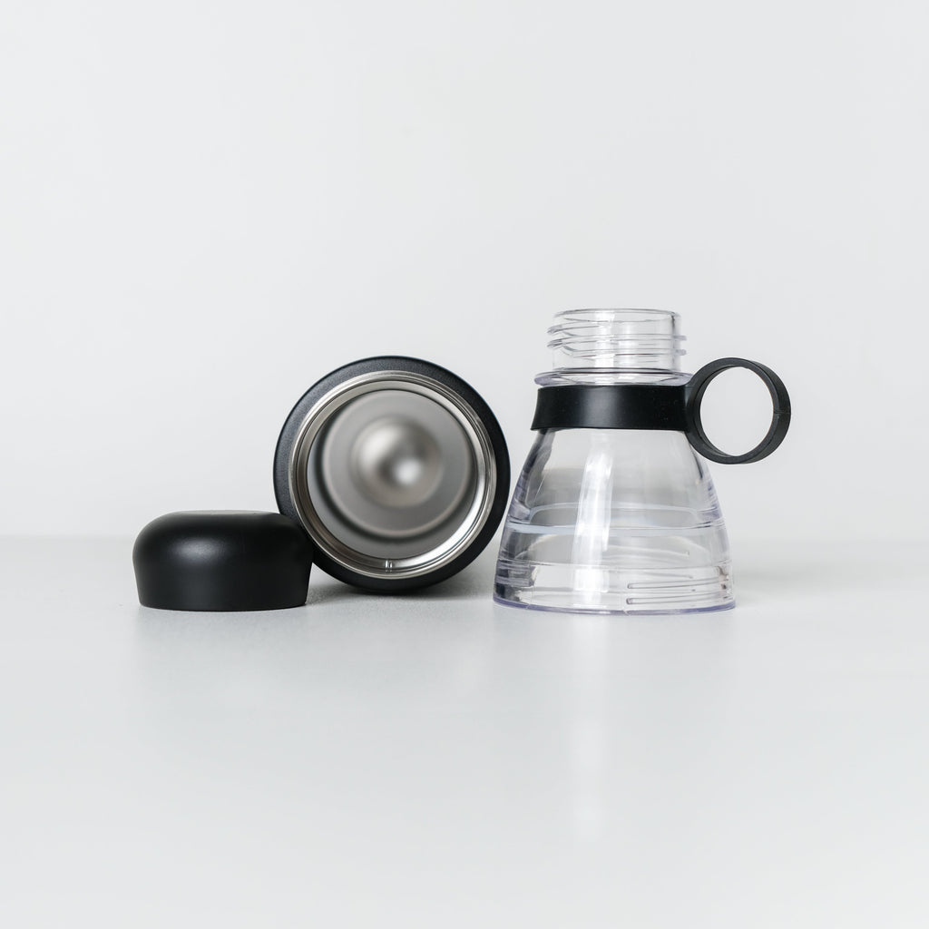 A clear plastic water bottle with a black handle and black collar, shown disassembled with its black cap and silver-toned inner lid placed beside it on a clean white studio background.