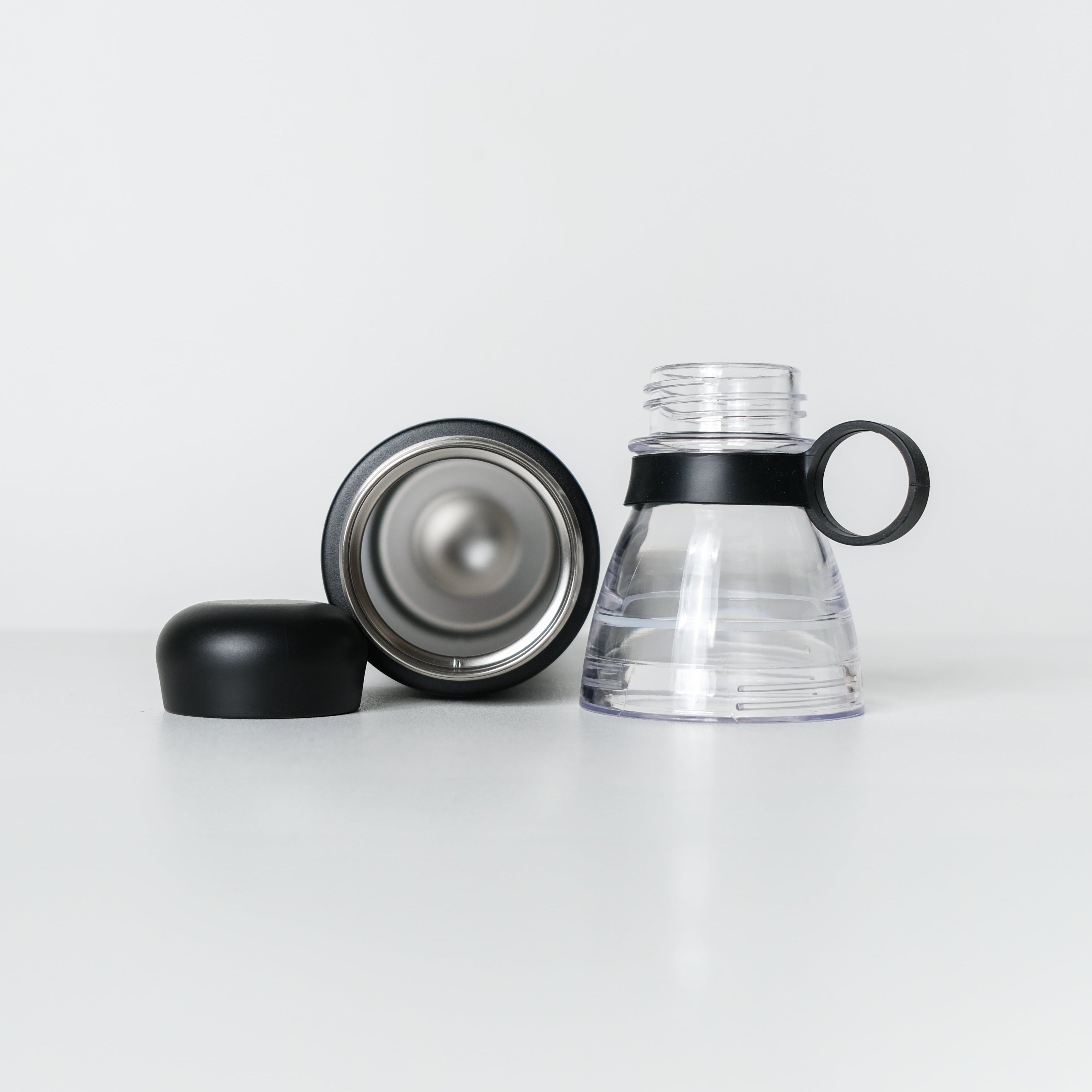 A clear plastic water bottle with a black handle and black collar, shown disassembled with its black cap and silver-toned inner lid placed beside it on a clean white studio background.