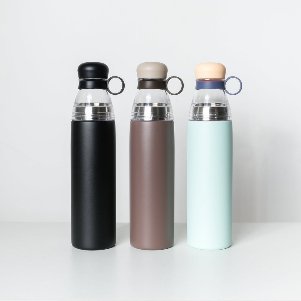 Three reusable insulated water bottles standing upright on a white shelf against a light gray wall. The bottles have matte black, taupe-brown, and pale mint-green bodies with clear upper sections and loop handles.