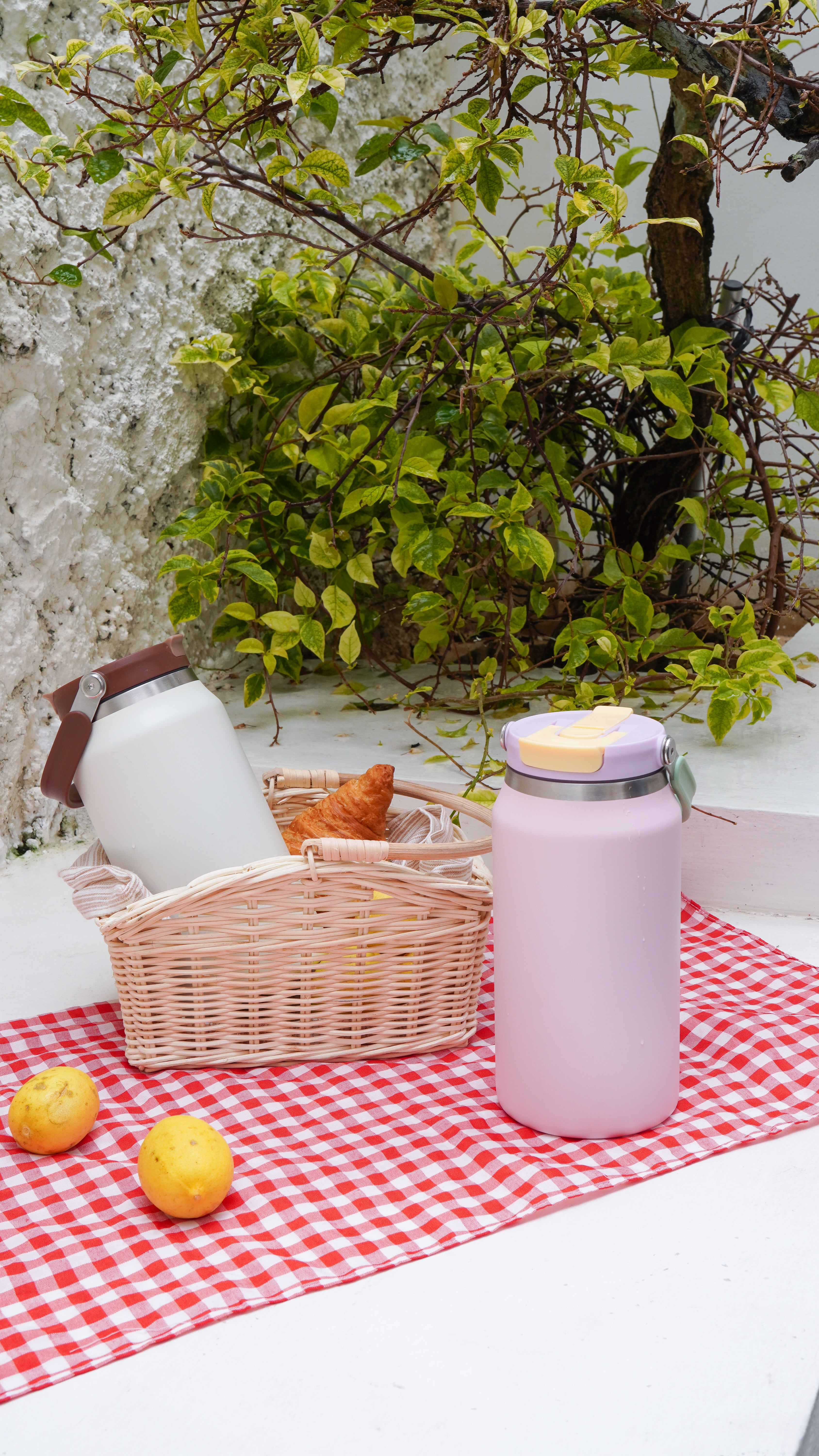 A pink insulated water bottle with a yellow lid sits on a red-and-white checkered picnic blanket beside a wicker basket holding a white bottle and a croissant, with lemons and greenery in the background.