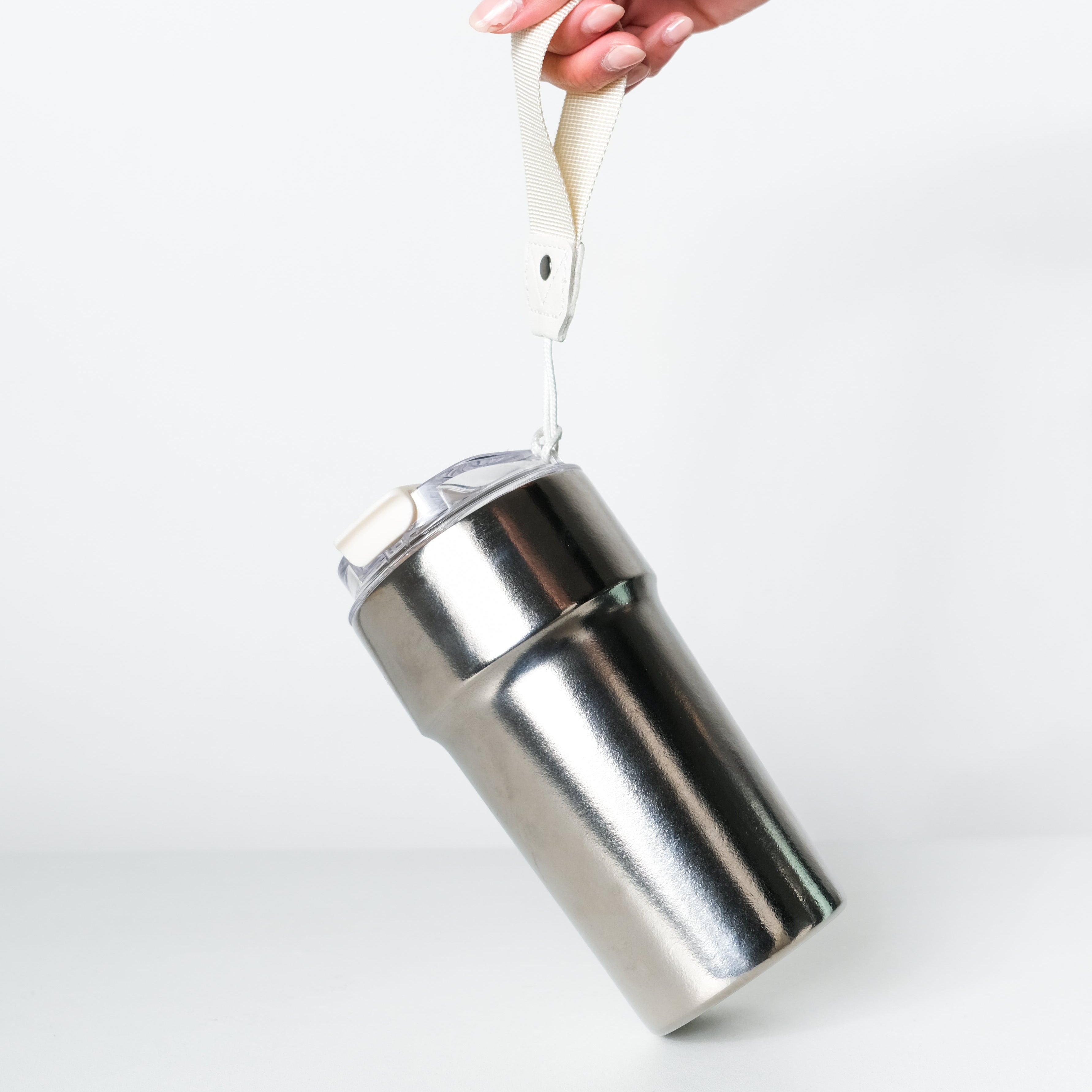 A stainless steel travel tumbler with a clear lid and a white carrying loop, held by a hand against a clean white background.