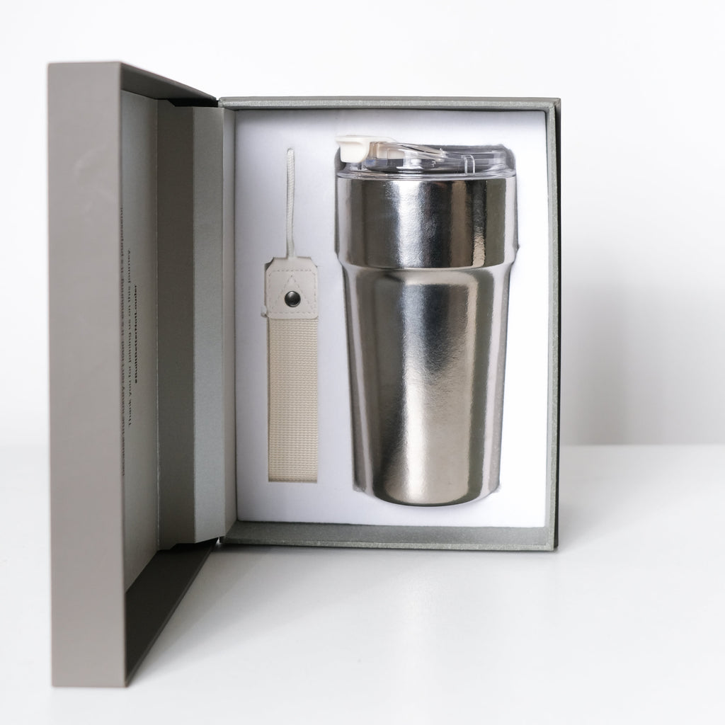 A stainless steel travel tumbler with a clear lid presented inside an open gray gift box, alongside a small cream-colored accessory, on a clean white surface against a white background.