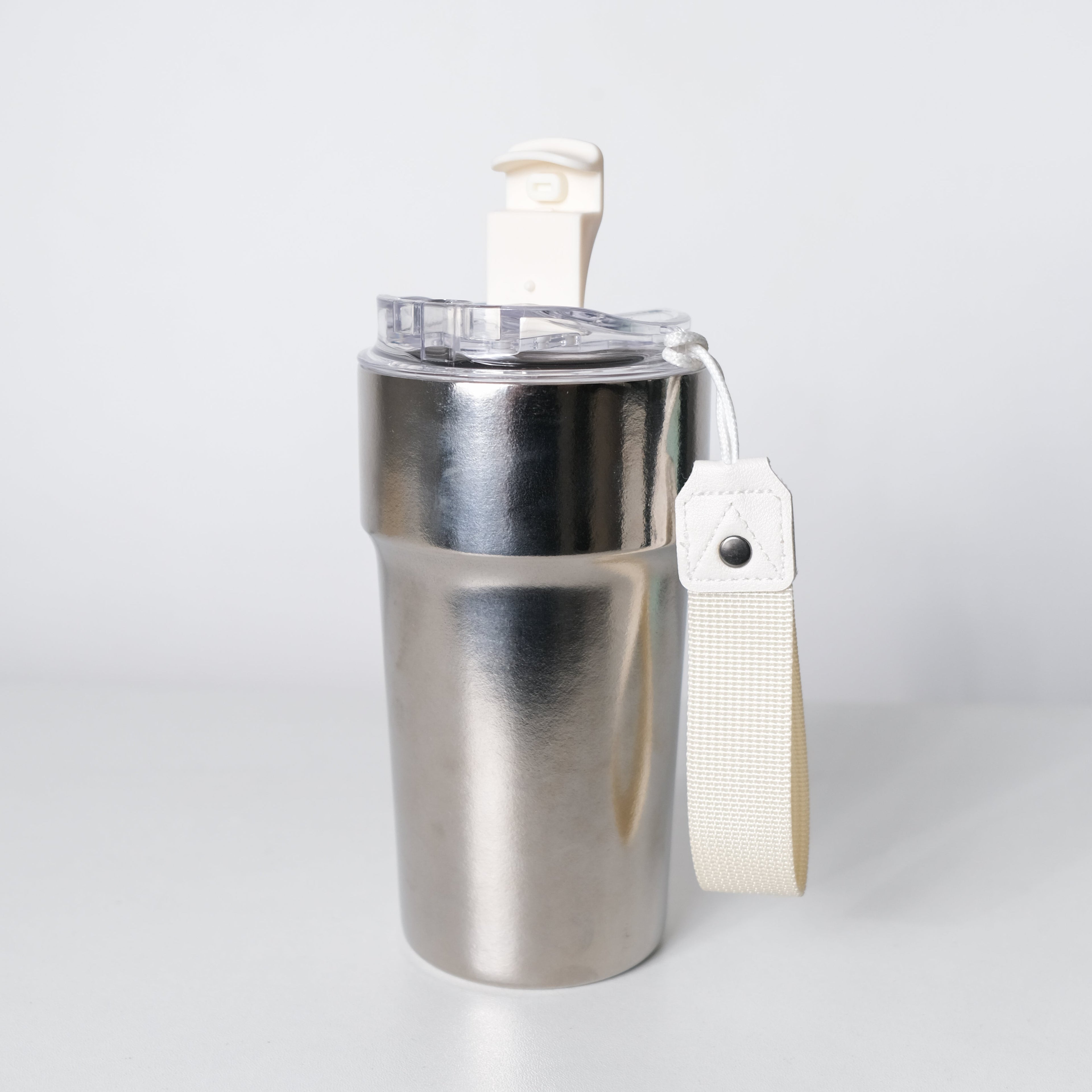 A stainless steel travel tumbler with a clear lid, cream-colored flip-top straw, and a matching fabric wrist strap, shown on a light gray background.