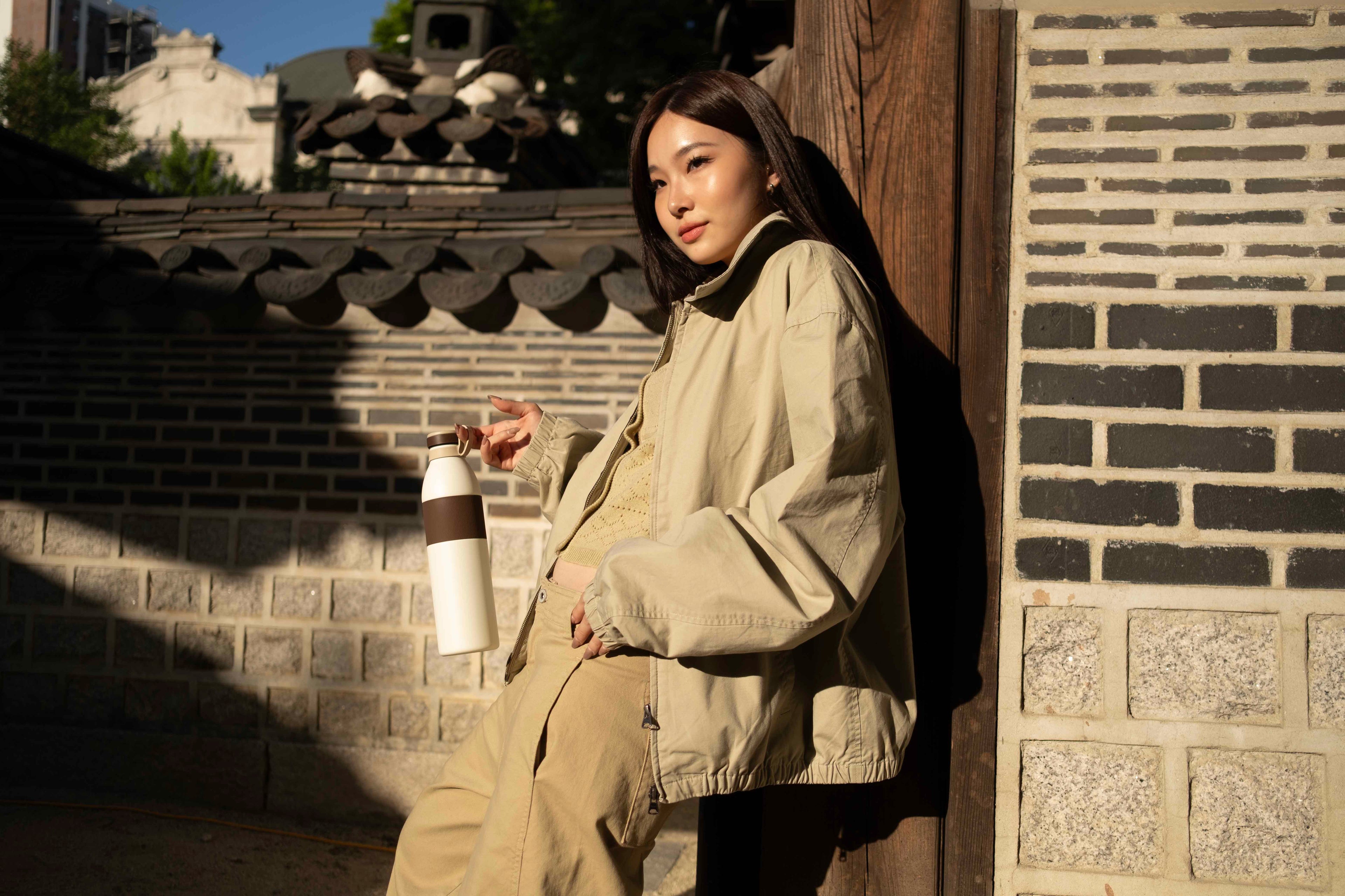 Woman in beige jacket sitting against a brick wall with a traditional building in the background